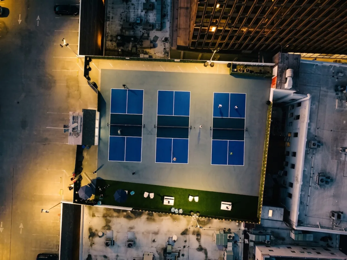 Straight-down drone view at dusk of the three blue rooftop courts and the turf lounge, lit for evening play